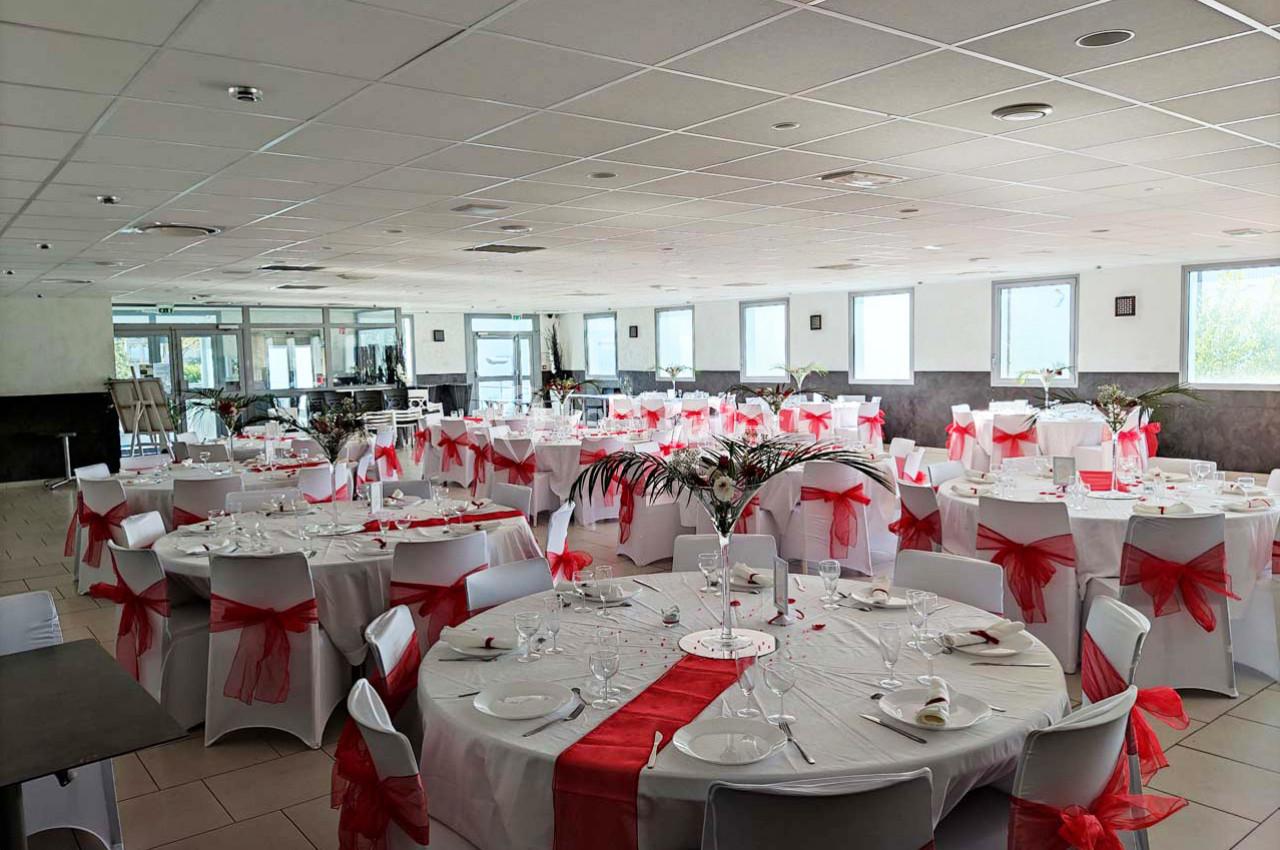 Salle de réception décorée avec des tables rondes, nappes blanches et nœuds rouges sur les chaises.