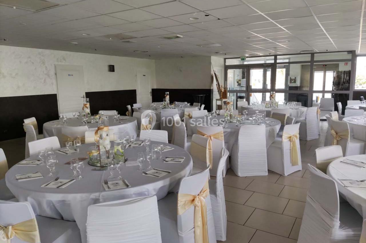 Salle de réception décorée avec des tables rondes, nappes blanches et chaises ornées de nœuds dorés.