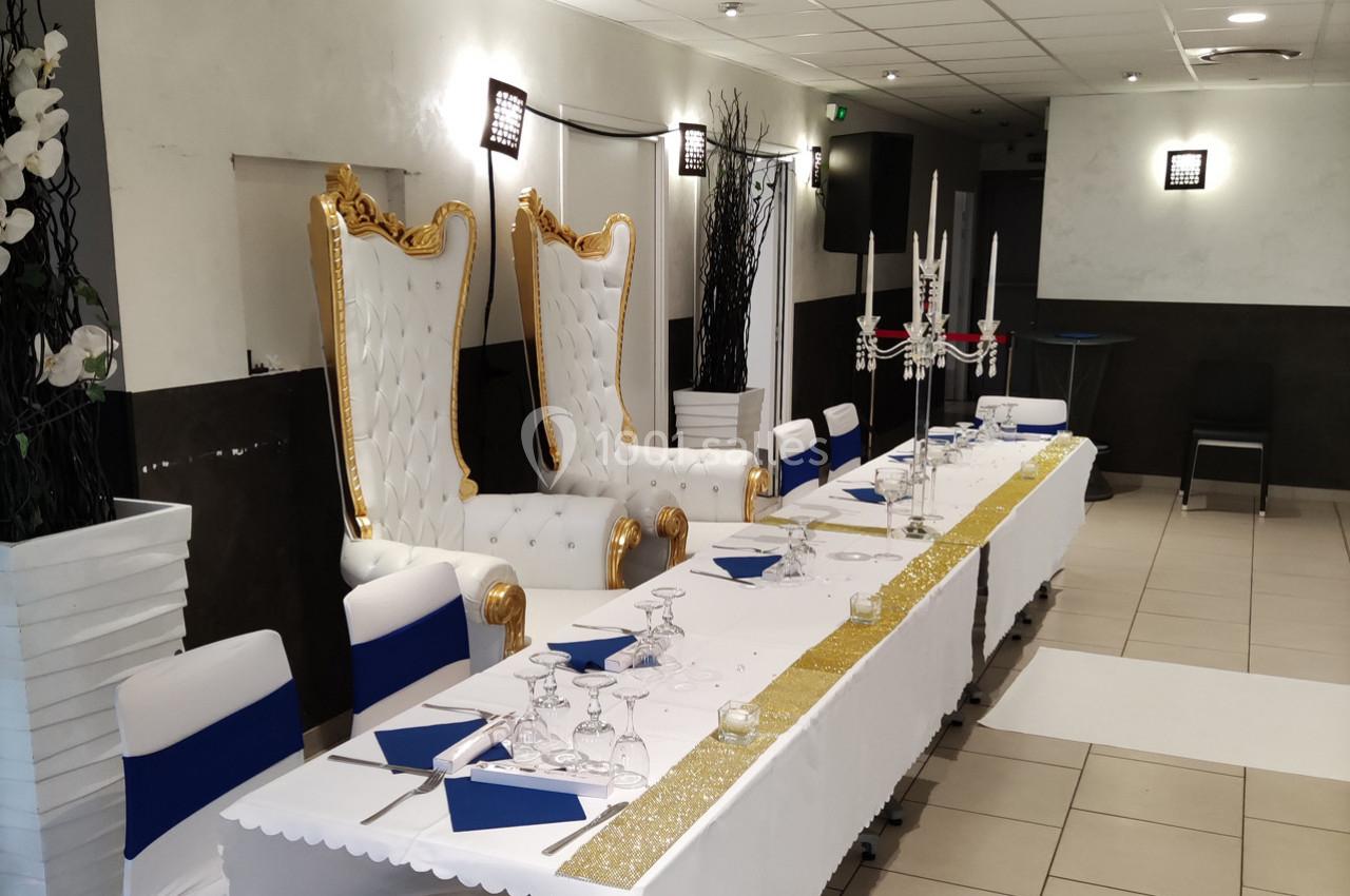 Salle de réception décorée avec une table longue, nappée de blanc et ornée de chandeliers, avec deux fauteuils dorés.