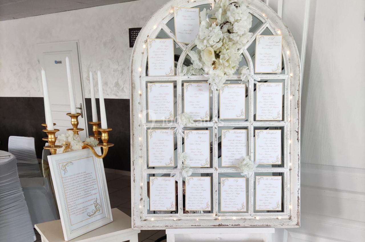 Plan de table de mariage affiché sur une fenêtre ancienne décorée de fleurs blanches, avec un chandelier doré à côté.