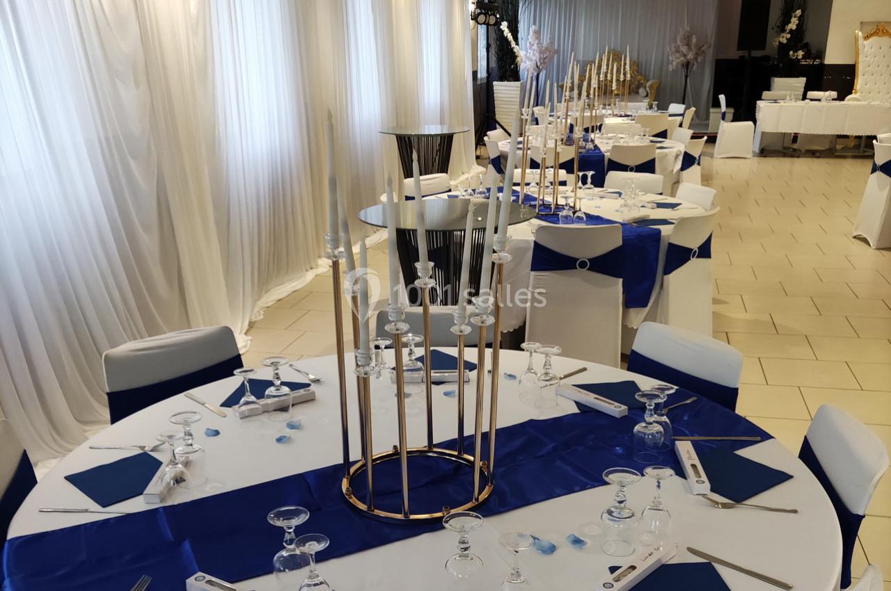 Salle de réception décorée avec des tables rondes, nappes blanches et bleues, et chandeliers dorés au centre.