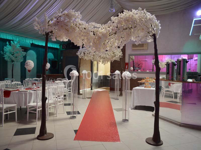 Salle de réception décorée avec des arbres artificiels fleuris, un tapis rouge et des tables dressées.