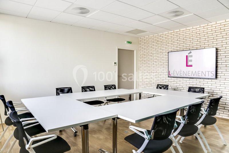 Salle de réunion lumineuse avec tables disposées en U, chaises noires et écran affichant un logo sur un mur en briques.