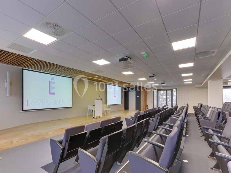 Salle de conférence moderne avec rangées de sièges, écran de projection et pupitre, éclairée par des plafonniers.