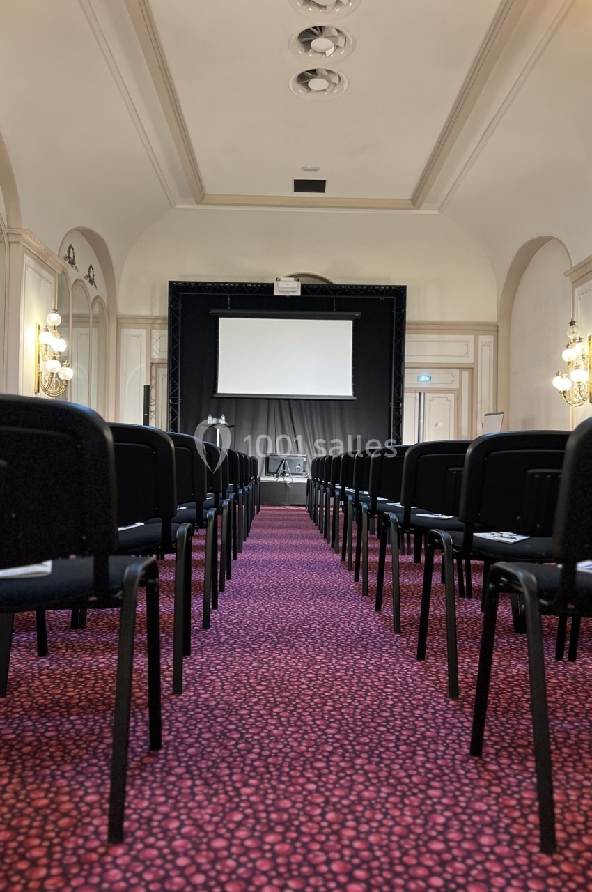 Salle de conférence avec rangées de chaises noires, tapis rouge à motifs et écran de projection au fond.