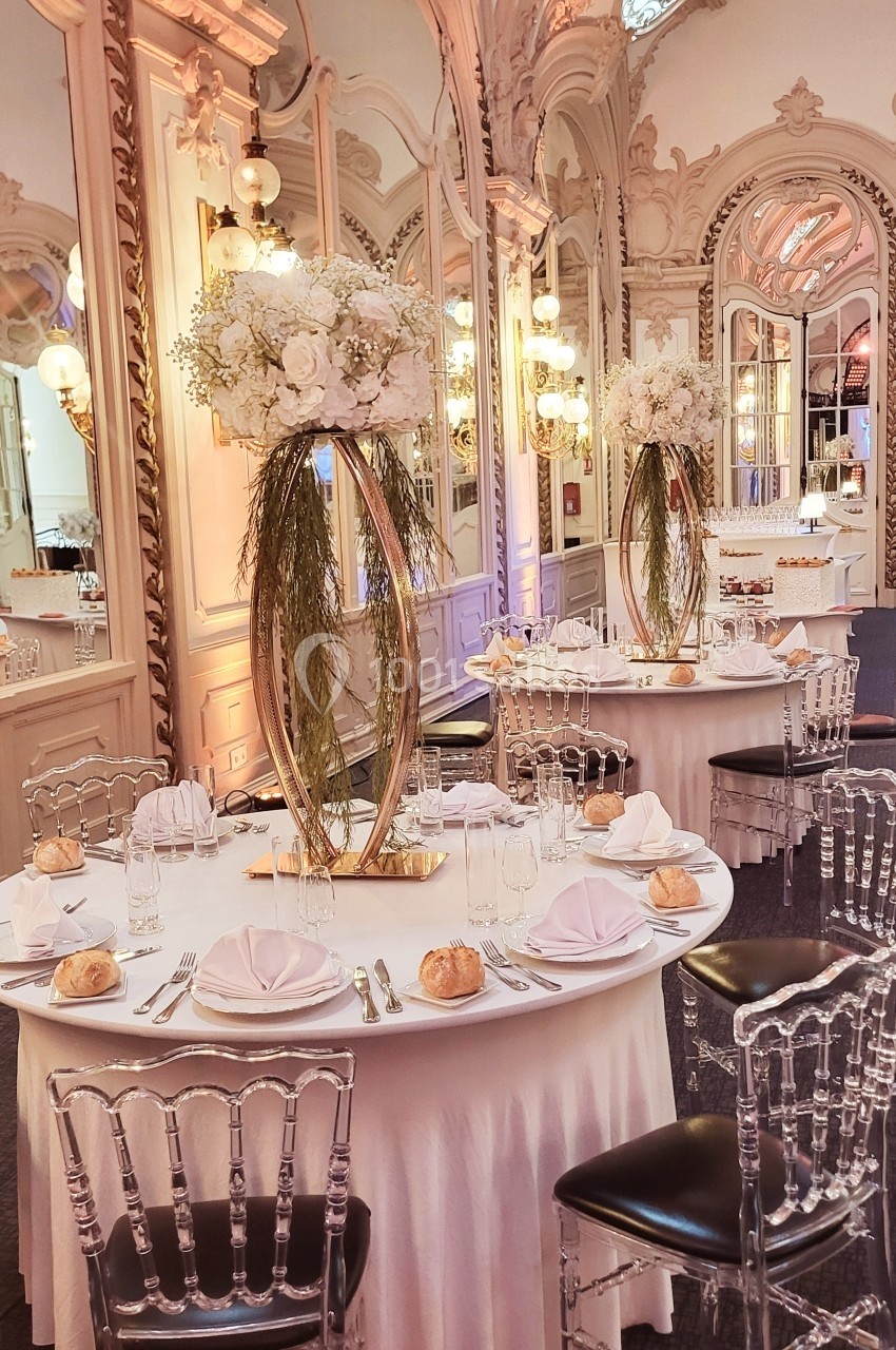 Salle de réception élégante avec tables rondes dressées, chaises transparentes et centres de table floraux.