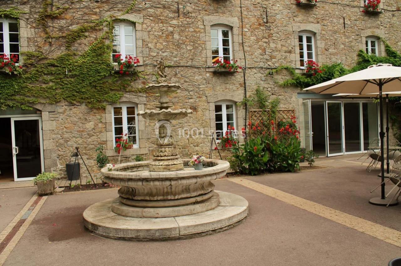 Cour intérieure avec une fontaine en pierre au centre, entourée de murs en pierre avec des fenêtres et des fleurs.