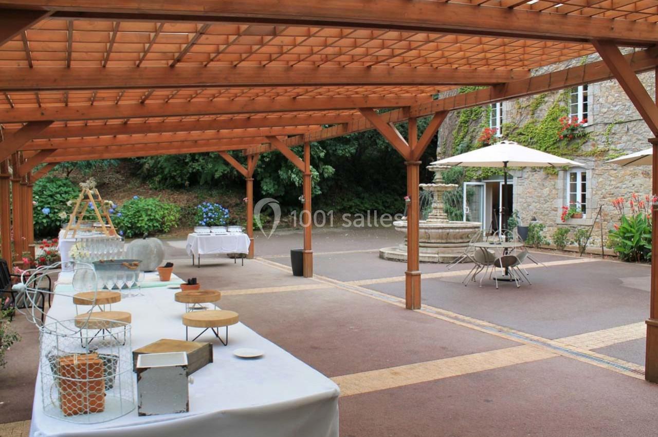 Espace extérieur avec pergola en bois, tables dressées, fontaine en pierre et végétation en arrière-plan.