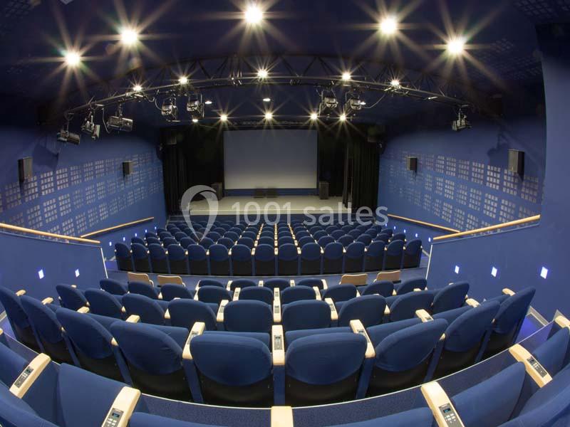 Salle de cinéma vide avec des rangées de sièges bleus, un écran blanc et des projecteurs allumés.