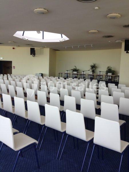 Salle de conférence vide avec des rangées de chaises blanches alignées et une estrade équipée de tables et de micros.