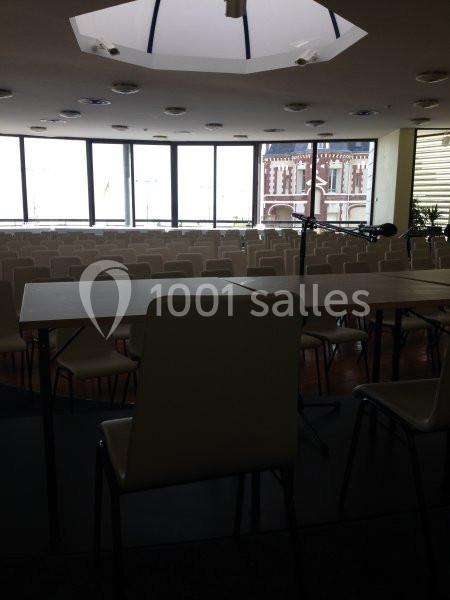 Salle de conférence vide avec des rangées de chaises blanches, une table et un micro, éclairée par une verrière.