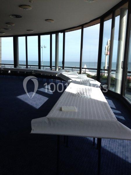 Salle lumineuse avec de grandes baies vitrées donnant sur la mer, tables alignées recouvertes de nappes blanches.