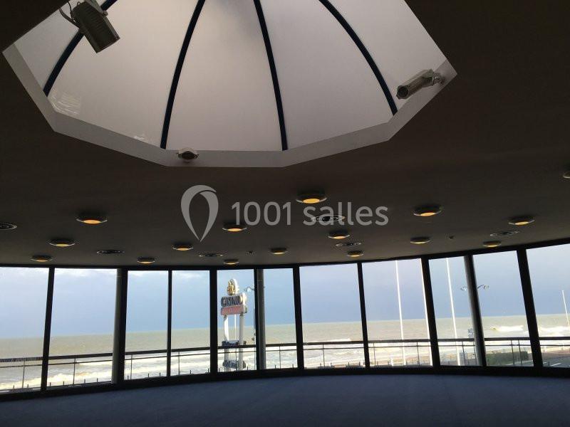 Vue intérieure d'une salle circulaire avec de grandes baies vitrées donnant sur la mer et un ciel partiellement nuageux.
