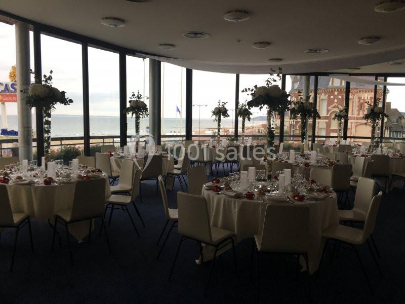 Salle de réception élégante avec tables rondes dressées, grandes baies vitrées offrant une vue sur la mer et des bâtiments.