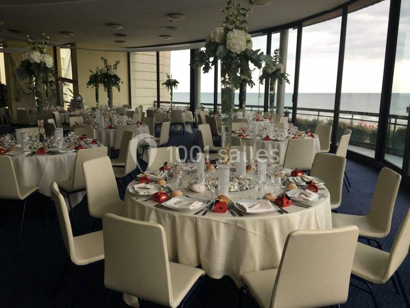 Salle de réception élégante avec tables rondes dressées, décor floral et vue sur la mer à travers de grandes baies vitrées.