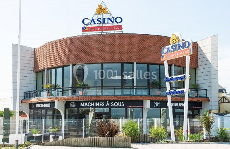 Façade d'un casino avec enseignes lumineuses, grandes baies vitrées et entrée principale entourée de plantes.