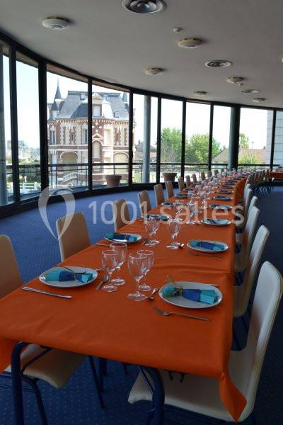 Salle avec de grandes baies vitrées, tables dressées avec nappes orange, assiettes bleues et verres, vue sur un bâtiment…