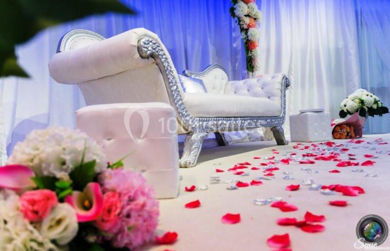 Canapé blanc orné d'argent sur une scène décorée de fleurs et de pétales rouges, éclairée par une lumière douce.