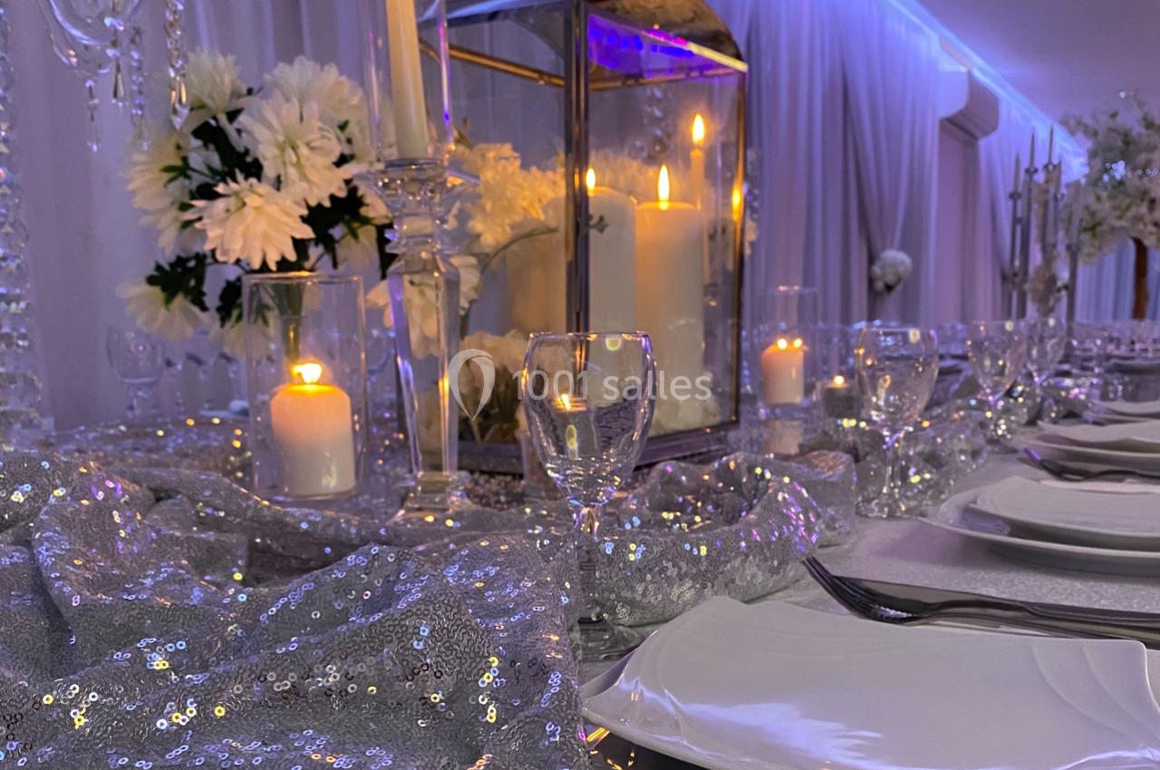 Table élégamment dressée avec nappes scintillantes, bougies allumées, fleurs blanches et vaisselle raffinée.