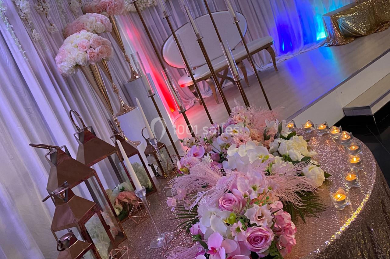 Décoration de salle avec table ornée de fleurs roses, lanternes cuivrées et bougies, devant une scène éclairée.