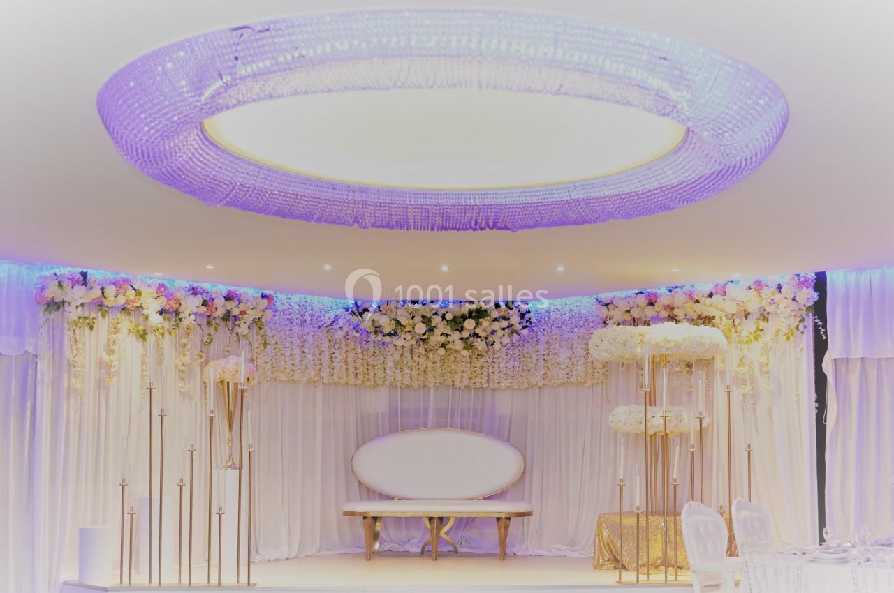 Décor de salle de réception avec un canapé blanc, rideaux et guirlandes de fleurs blanches sous un lustre circulaire…