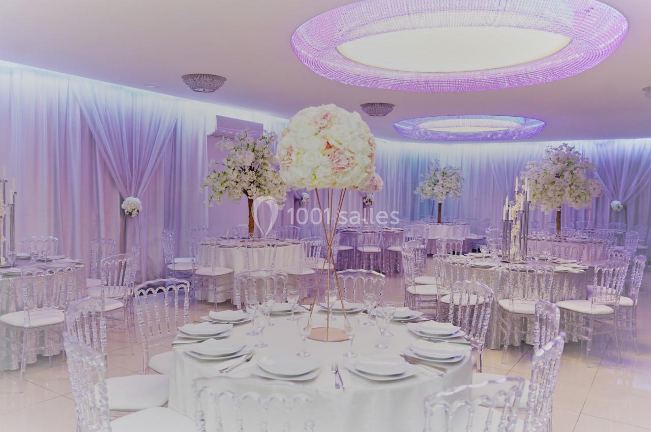Salle de réception élégante avec tables rondes dressées, chaises transparentes et décorations florales blanches.
