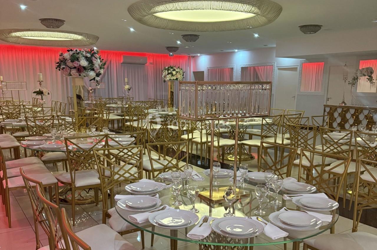 Salle de réception élégante avec tables rondes dressées, chaises dorées et décorations florales sous un éclairage tamisé.