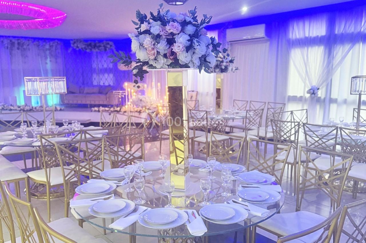 Salle de réception élégante avec tables rondes dressées, décor floral central et éclairage bleu tamisé.
