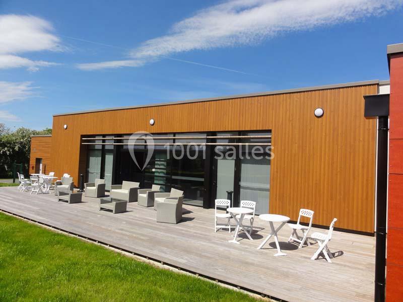 Bâtiment moderne avec façade en bois, grande terrasse en bois aménagée de tables et chaises, sous un ciel dégagé.