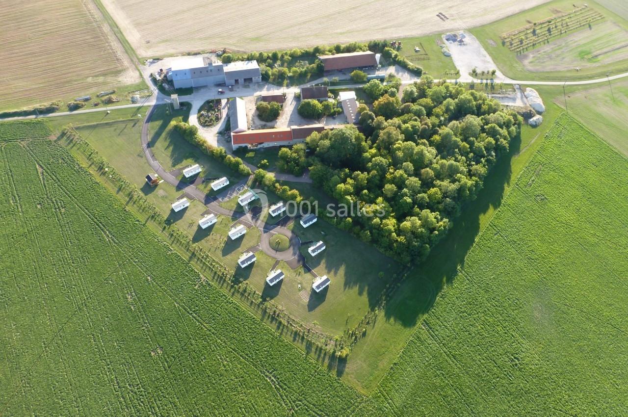 Vue aérienne d'un complexe avec bâtiments, espaces verts, arbres et structures dispersées au milieu de champs agricoles.