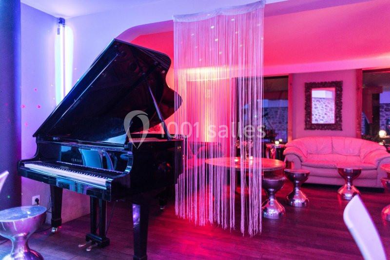 Piano à queue noir dans un salon éclairé par des lumières roses, avec canapé et tables en arrière-plan.