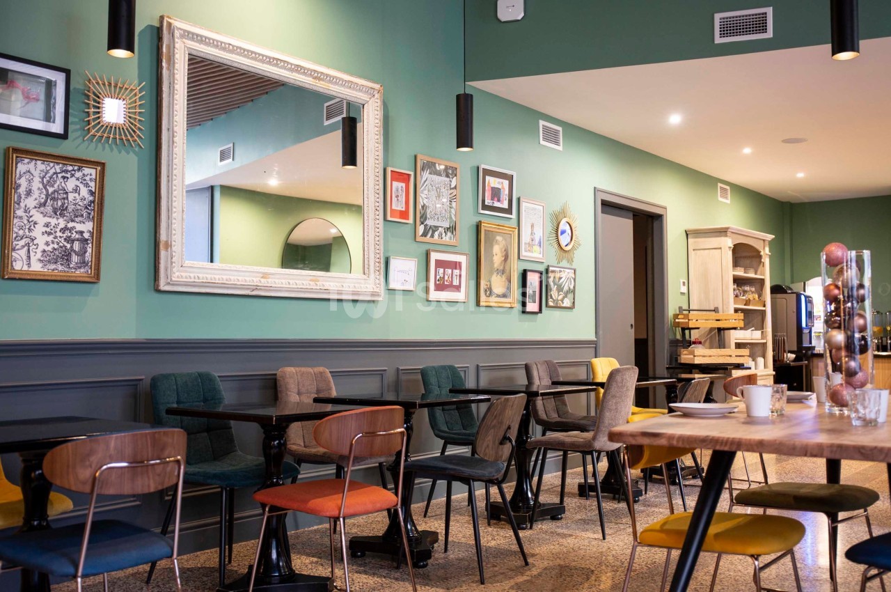 Salle de restaurant moderne avec murs verts, tables en bois, chaises colorées et décorations murales variées.