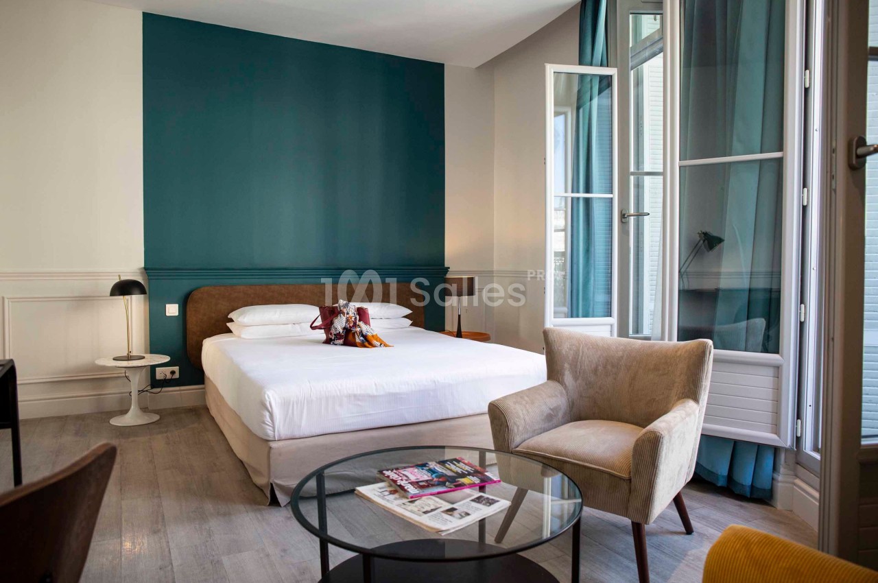 Chambre lumineuse avec lit double, fauteuil, table basse, magazines et grande fenêtre ouverte sur des rideaux bleus.