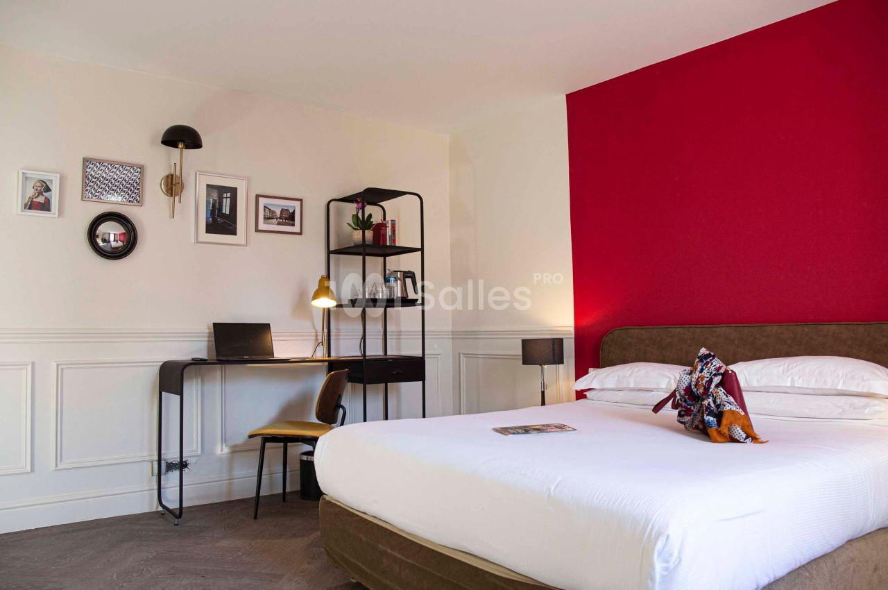 Chambre lumineuse avec un lit double, un bureau équipé d'un ordinateur portable et un mur rouge en arrière-plan.