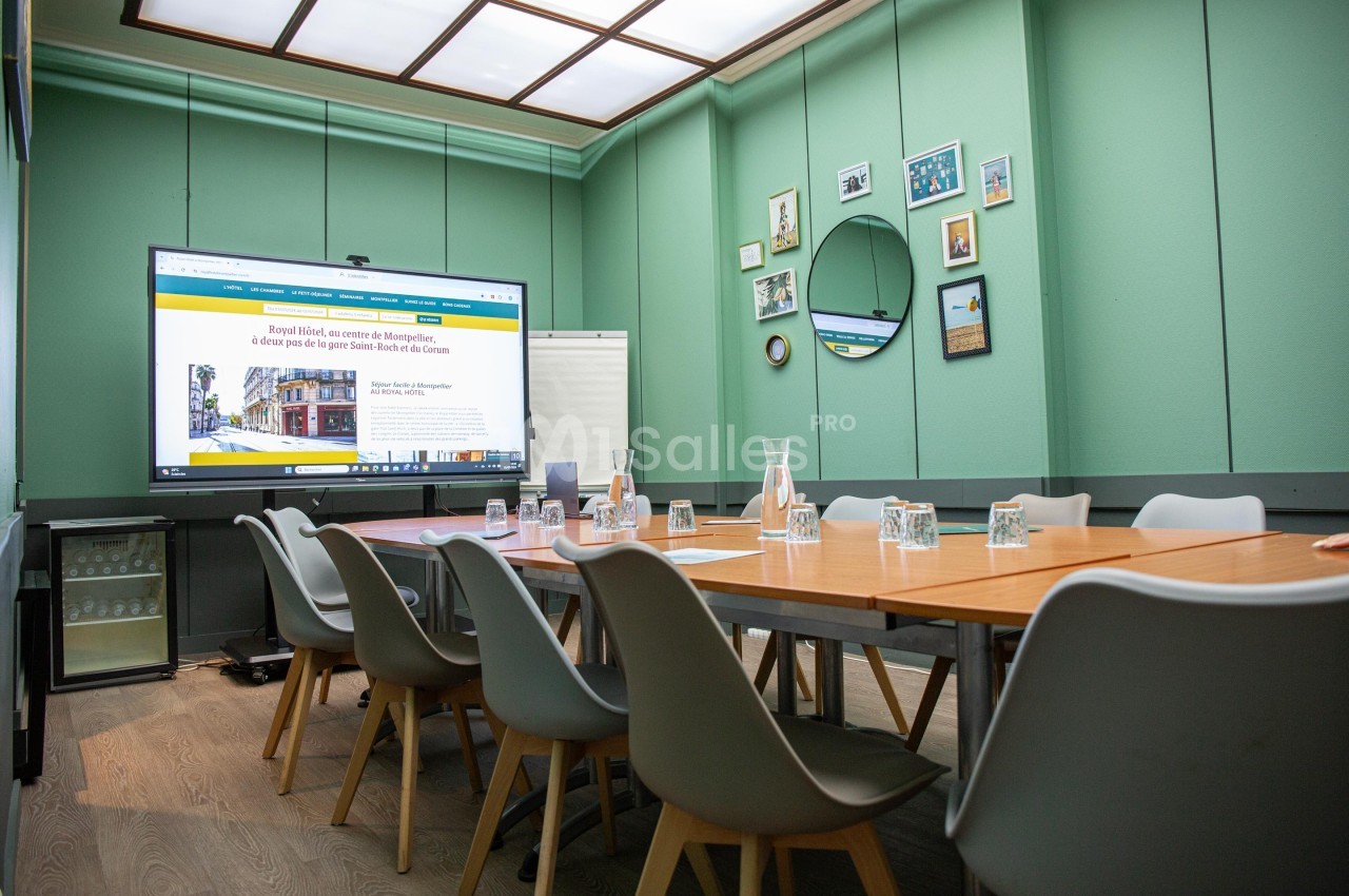 Salle de réunion moderne avec table en bois, chaises grises, écran allumé, murs verts et décorations murales.