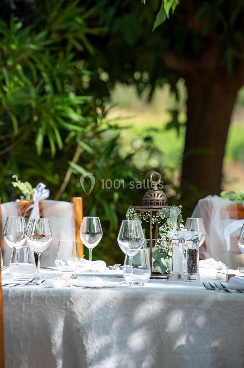 Table dressée en extérieur avec nappes blanches, verres, lanternes décoratives et végétation en arrière-plan.