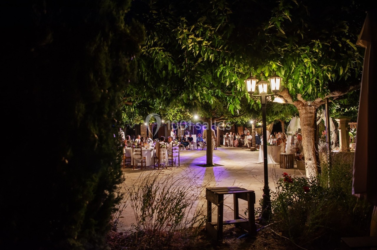 Vue nocturne d'une terrasse éclairée avec des tables dressées et des invités réunis sous des arbres.