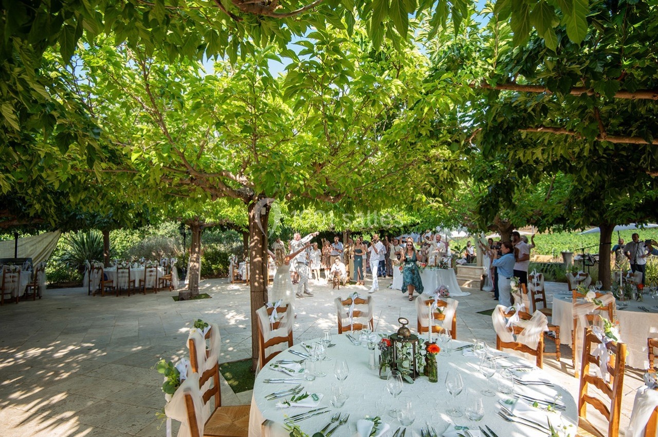 Tables décorées pour un événement en plein air sous des arbres, avec des invités en arrière-plan.