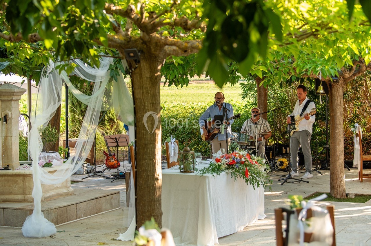 Groupe de musiciens jouant en plein air sous des arbres, près d'une table décorée et d'une fontaine.