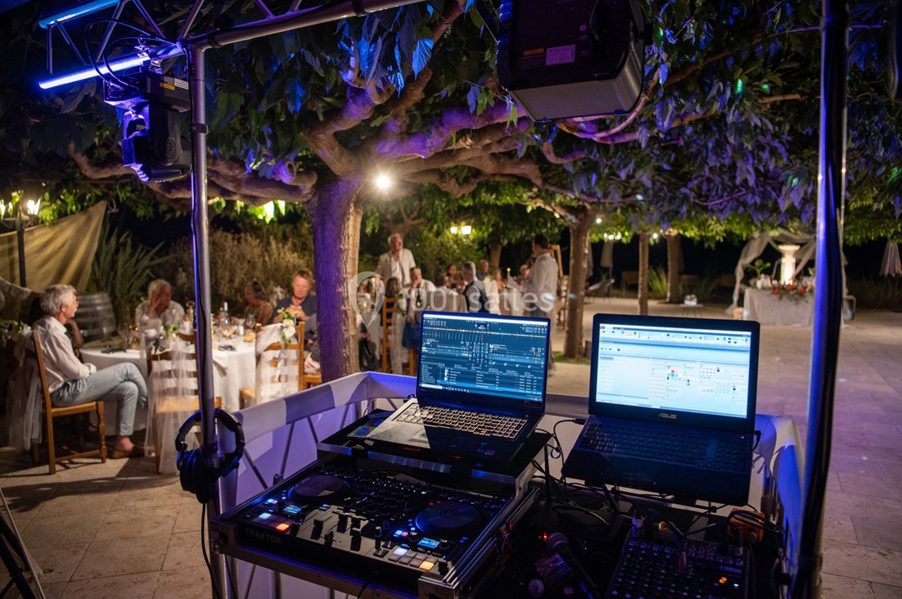Poste de DJ avec ordinateurs et matériel audio lors d'une soirée en extérieur sous des arbres illuminés.