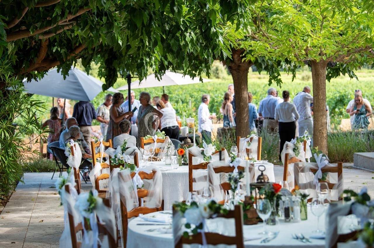 Tables décorées pour un événement en plein air sous des arbres, avec des invités discutant en arrière-plan.