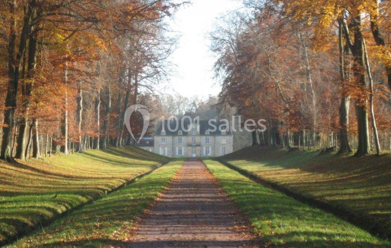 Allée bordée d'arbres aux feuilles d'automne menant à un manoir en pierre sous un ciel clair.