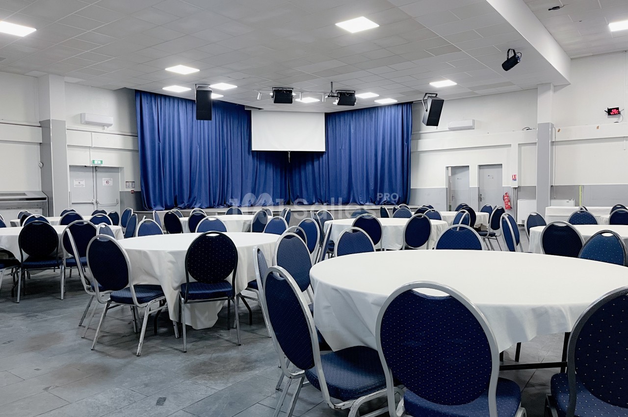 Salle de réception avec des tables rondes dressées de nappes blanches et une scène équipée de rideaux bleus.