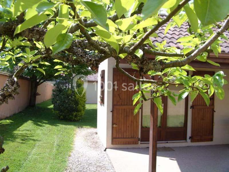 Location salle Saint-Georges-d'Espéranche (Isère) - Les Jardins du Castel #18 Maison avec volets en bois, entourée d'un jardin verdoyant et vue partielle d'un arbre au premier plan.