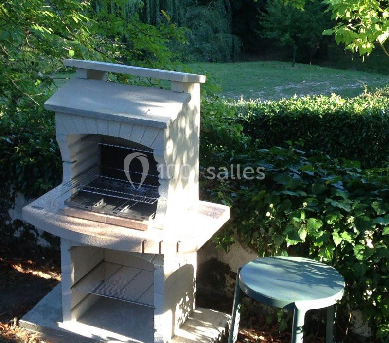 Location salle Saint-Georges-d'Espéranche (Isère) - Les Jardins du Castel #12 Barbecue en pierre dans un jardin verdoyant, accompagné d'un tabouret vert et entouré de haies et d'arbres.