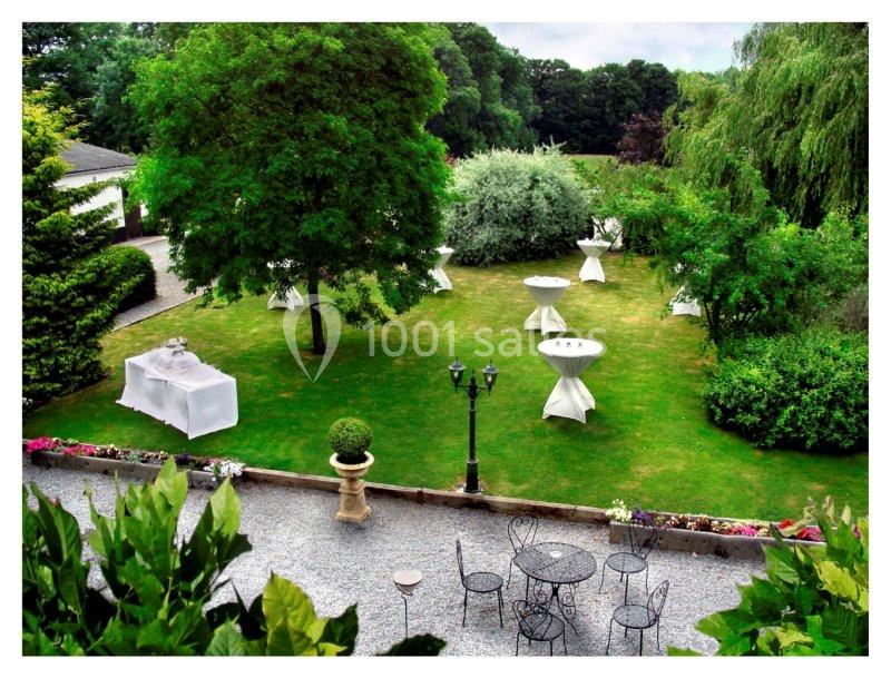 Jardin verdoyant aménagé pour un événement avec tables hautes nappées, chaises en fer forgé et allée gravillonnée.