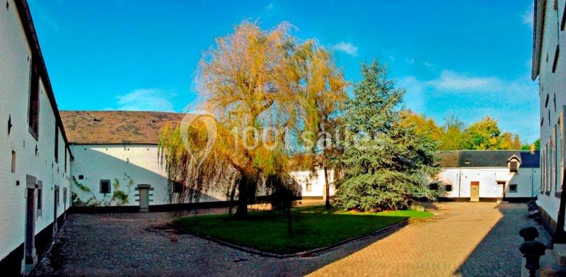 Cour intérieure pavée entourée de bâtiments blancs, avec un arbre au centre et un ciel bleu dégagé.