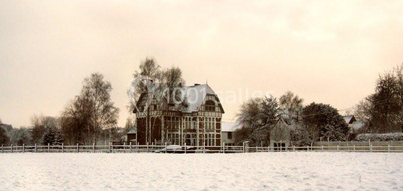 Maison à colombages entourée d'arbres et d'une clôture, dans un paysage enneigé sous un ciel nuageux.