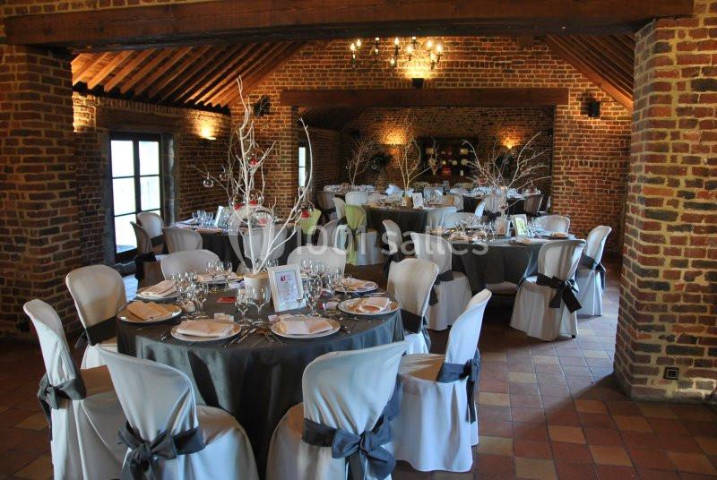 Salle de réception en briques avec tables rondes décorées, nappes grises et chaises blanches ornées de nœuds noirs.