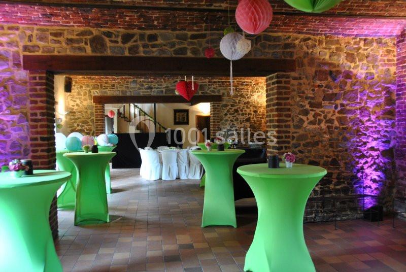 Salle en pierre décorée pour un événement, avec tables hautes couvertes de nappes vertes et suspensions colorées.